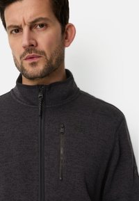 Dunkelgraue Fleecejacke mit hohem Kragen, Brusttasche mit Reißverschluss und strukturierter Oberfläche. Reißverschluss-Details in schwarzem Kontrast.