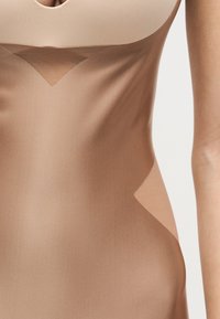 Κοντινό πλάνο ενός μπεζ εφαρμοστού φορέματος με διαφανή τούλινα πάνελ στο στήθος και τις πλευρές, που φοριέται από ένα άτομο σε απλό φόντο.