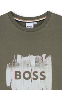 Oliwkowa koszulka crewneck z białym wzorem pędzla i miejskim pejzażem za wyraźnym napisem "BOSS" na przodzie.