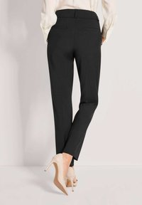 Schwarze maßgeschneiderte Hosen mit geradem Schnitt, ausgestattet mit einer glatten Textur und zwei Gesäßtaschen. Kombiniert mit hautfarbenen High Heels.