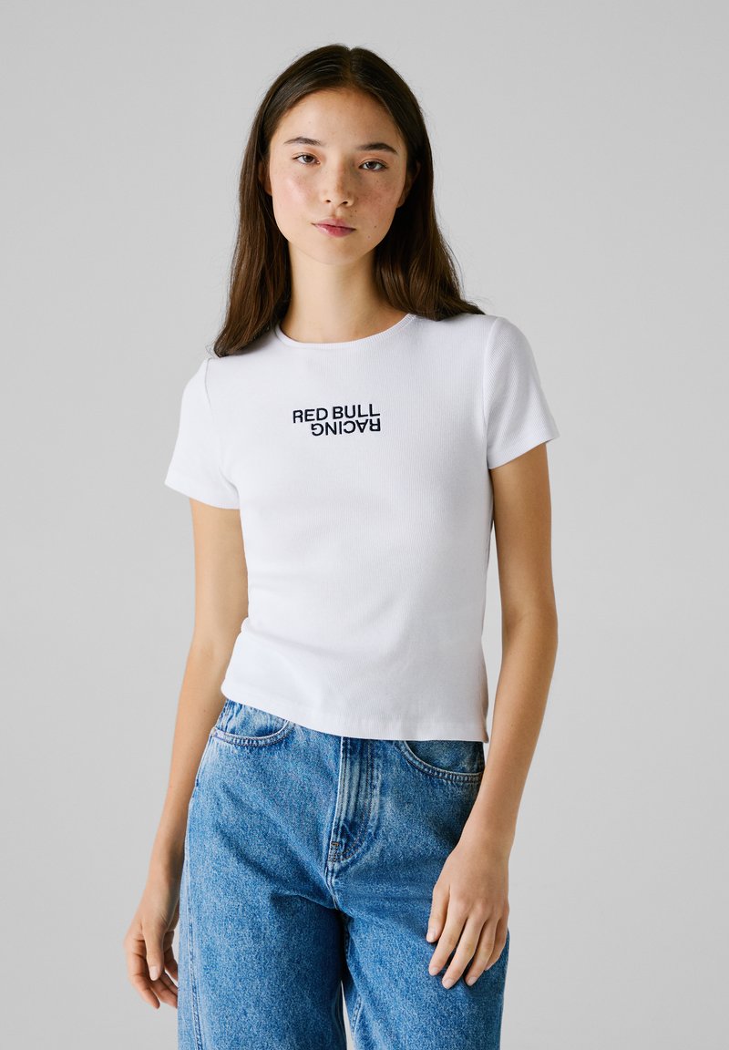 Hvid kortærmet T-shirt med ribbet tekstur og sort "RED BULL RACING" grafik trykt på fronten, parret med blå højtaljede jeans.