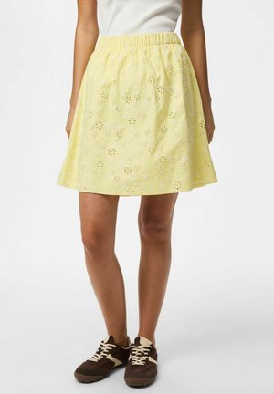 Žena má na sebe žltú kvetinovú A-čkovú sukňu s dierkami, biely tielko a hnedé a béžové tenisky, stojí na bielom pozadí.