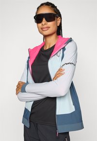 Hele sinine spordik jacket roosa fliisvoodriga kapuutsiga, must meshtopp all, ja julged sinised aktsendid. Stiilsed prillid täiendavad välimust.