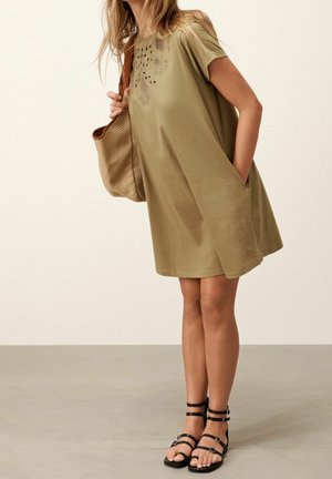 Femme portant une robe olive à manches courtes avec des poches, des sandales noires à brides et un grand sac d'épaule tissé.