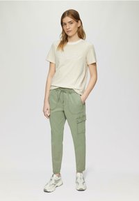 Hellbeige gestreiftes T-Shirt mit dem Schriftzug "shine", kombiniert mit hellgrünen Cargo-Hosen, die seitliche Taschen und einen Kordelzugbund haben. Silberne Sneaker.