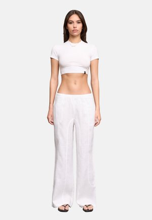 Junge Frau mit langen dunklen Haaren, die ein weißes kurzes T-Shirt, lockere weiße Hose und schwarze Flip-Flops trägt, vor einem einfarbigen Hintergrund stehend.