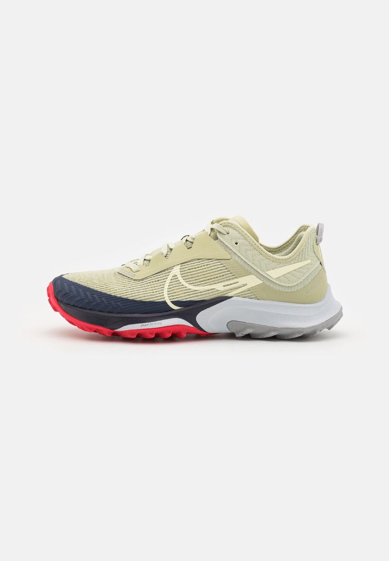 Chaussure de sport beige avec un design à lacets, semelle texturée noire et rouge, et un logo Nike swoosh jaune clair sur le côté.