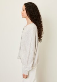 Světle šedý vlněný cardigan s volným střihem, kulatým výstřihem, žebrovanými manžetami a zapínáním na knoflík. Měkká textura s minimálními detaily.