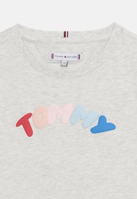 Ανοιχτό γκρι μπλουζάκι με στρογγυλή λαιμόκοψη και πολύχρωμο, φουσκωτό σχέδιο κειμένου "Tommy" στο στήθος, με ετικέτα Tommy Hilfiger μέσα στο γιακά.