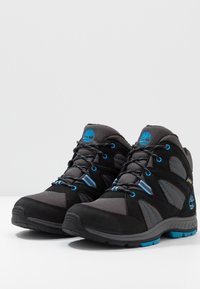 Zwarte en grijze wandelschoenen van mesh en suede materialen, met blauwe accenten en rubberen zolen. Voorzien van een vetersysteem en logo-detail.