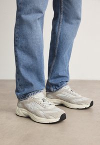 Osoba v lehce šedých a béžových šněrovacích teniskách s tlustými podrážkami a modrých džínách stojí na hladké podlaze.