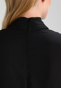 Svart blus med hög krage, subtil rynkning och dold dragkedja bak för stängning. Slät tygstruktur.