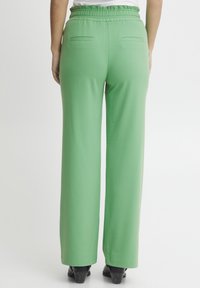 Osoba w luźnych, jasnoniebieskich spodniach z wysokim stanem i czarnych butach, stoi na tle jednolitego białego tła.