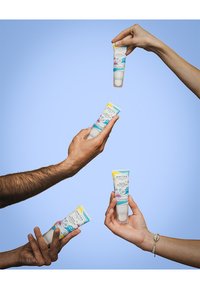 Solkremtuber holdt av hender i forskjellige hudtoner. Tubene har klare etiketter som fremhever SPF 50, samt fargerike bølgemønstre.