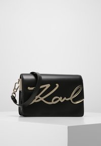 Černá kožená kabelka se zlatým nápisem "Karl" na přední straně, s odepínacím páskem a elegantním obdélníkovým tvarem.
