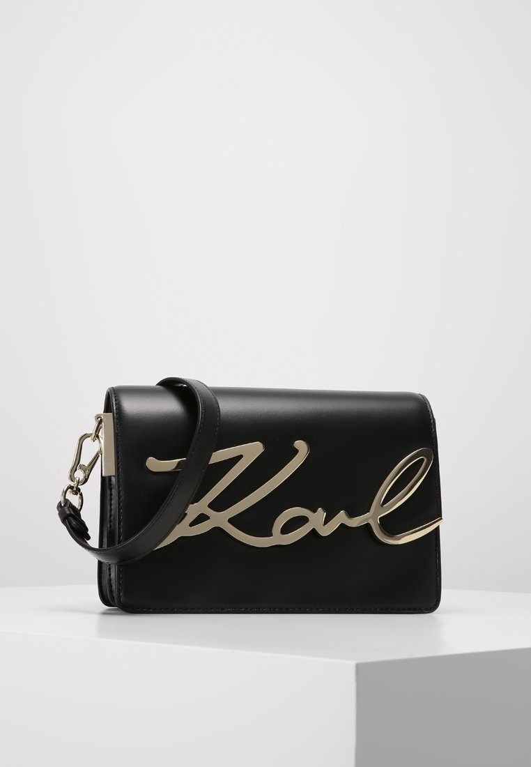 Černá kožená kabelka se zlatým nápisem "Karl" na přední straně, s odepínacím páskem a elegantním obdélníkovým tvarem.