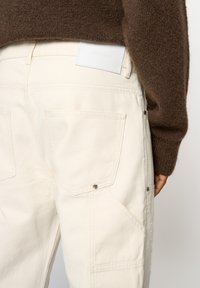 Personne portant un pantalon en denim écru avec des poches à l'arrière et sur les côtés, une étiquette blanche sur la ceinture, et un pull brun en matière duveteuse.