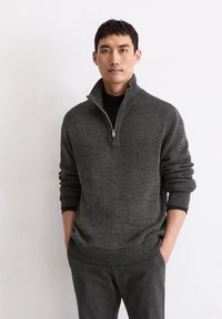 Grauer gerippter Pullover mit einem halben Reißverschluss am Kragen und umgeschlagenen Bündchen, aus strukturiertem Strickmaterial, kombiniert mit dunkelgrauen, vorn flachen Hosen.