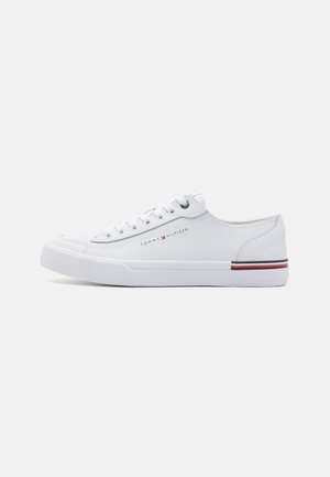 Zapatillas blancas de cuero con perfil bajo, punta redonda y suela plana de goma. Presenta ojales plateados y un acento a rayas cerca de la suela.