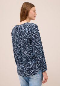 Mujer con cabello castaño lacio que lleva una blusa suelta azul marino con manchas irregulares de color azul claro y jeans azul claro, mirando de lado.
