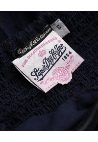 Námořnická modrá tkanina se shrnutou strukturou; obsahuje růžovou etiketu s nápisem "SuperDry Co. Japan" a "1954" v dekorativním písmu. Velikost S je uvedena.