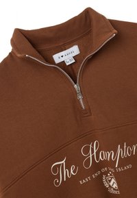 Felpa marrone con zip a un quarto, con la scritta "The Hamptons East End of Long Island" e un piccolo stemma sotto la zip.