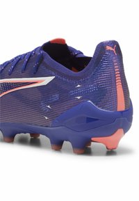 Fialové futbalové kopačky s textúrovaným vrchom, bielymi akcentmi a flexibilnou fialovou podrážkou s oranžovými detailmi a viditeľnými kolíkmi pre lepšiu trakciu.