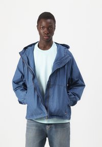 Lys blå denimjakke med hette, frontglidelås og elastiske mansjetter, lagd over en lys blå t-skjorte, kombinert med lyseblå jeans.