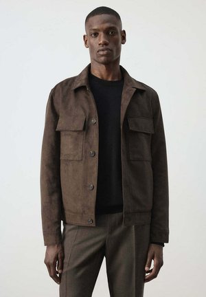 Hombre con chaqueta marrón oscuro abotonada sobre una camisa negra y pantalones marrones, de pie frente a un fondo claro y liso.