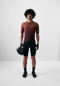 Îmbrăcăminte scurtă pentru ciclism cu un gradient de la maro la negru, mâneci scurte, fermoar frontal, completată cu șorturi negre și mănuși, ținând un cască neagră.