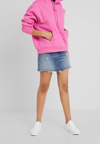 Sweat à capuche rose avec poche avant, associé à une mini-jupe en denim à ourlet effiloché et délavage clair. Des baskets blanches complètent la tenue.