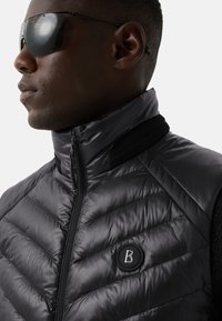 Zwart gewatteerd vest met een hoge kraag, voorzien van een doorgestikte textuur, ritssluiting en een rond logo-accent op de borst. Inclusief mesh details.