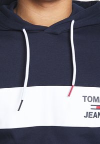 Granatowa i biała hoodie z białymi sznurkami ściągającymi, z napisem "TOMMY JEANS" oraz małym logo w kolorze czerwonym, białym i niebieskim.