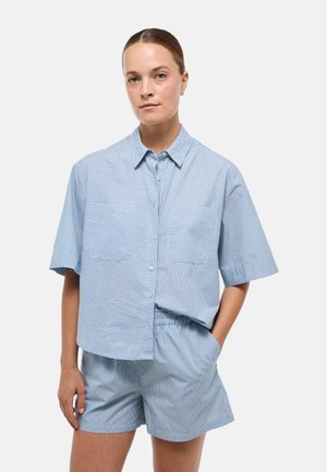 Jeune femme à la peau claire et aux cheveux attachés porte une chemise à manches courtes rayée bleue et un short assorti, debout avec une main dans la poche.