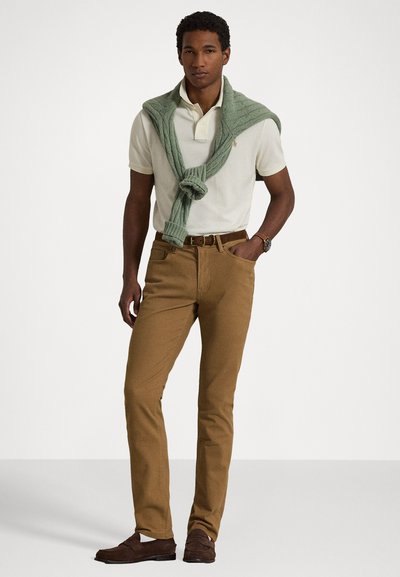 Polo krekls krēmkrāsā ar dekoratīvu apkakli, zaļš adīts džemperis, kas sasiets ap kaklu, brūni auduma bikses un tumši brūni mokasīni.