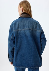 Jeansjacka i mörkblått med krage av brunt läder, med avslappnad passform och synliga sömnadsdetaljer. Visas från baksidan.