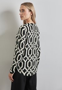 Het langemouwen shirt heeft een zwart-wit geometrisch patroon met hexagonale vormen. De stof is glad, met een ontspannen pasvorm en een ronde halslijn.