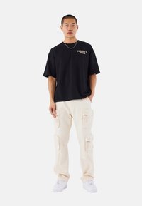 Camiseta negra de manga corta con el texto "Project Paris", combinada con pantalones cargo beige con múltiples bolsillos y cremalleras. Zapatillas blancas.