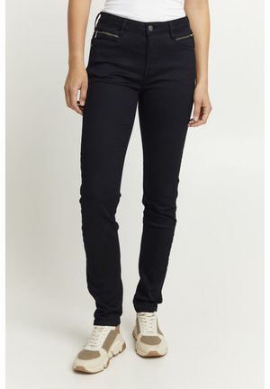 Zwarte skinny jeans van rekbare stof. Voorzien van een ritszakken aan de zijkant en een aangesloten ontwerp, gecombineerd met lichtgekleurde sportschoenen.