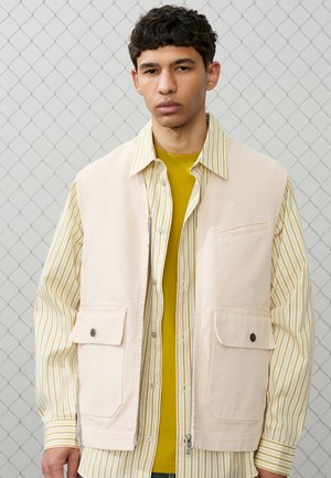 Tânăr purtând o vestă utilitară bej peste un tricou cu dungi galbene și un tricou muștar, stând în fața unui gard din plasă metalică.