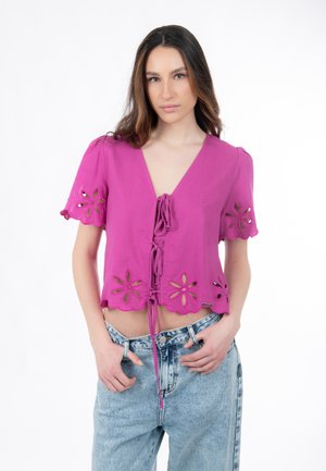 Kurze pinke Bluse mit kurzen Puffärmeln, Schnürung vorne und floralen Cut-out-Mustern am Saum. Kombiniert mit hoch taillierten Jeans.