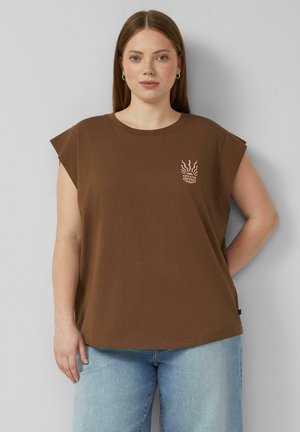 Jeune femme debout contre un mur gris portant un débardeur marron avec un petit motif de soleil et un jean bleu clair.