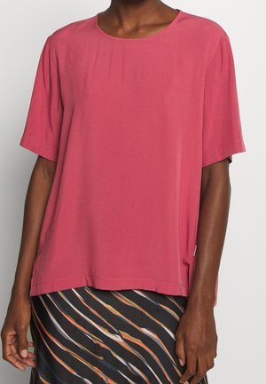 Persoon die een loszittende roze top met korte mouwen draagt en een zwarte rok met oranje, witte en gele diagonale strepen.