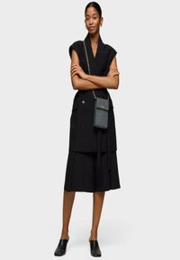 Vestido negro de doble botonadura con falda de capas, mangas cortas y cinturón en la cintura. Combinado con un pequeño bolso cruzado y mules negros.