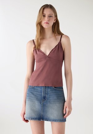 Giovane donna che indossa un top senza maniche color malva con rifiniture in pizzo e una minigonna di jeans blu su uno sfondo chiaro e uniforme.