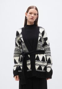 Πλεκτή ζακέτα με γεωμετρικά μοτίβα σε μαύρο και λευκό, ανοιχτό μπροστινό μέρος και μαύρη ζώνη που δένει. Μακριά μανίκια με τελειώματα σε μανσέτα, απαλή υφή.