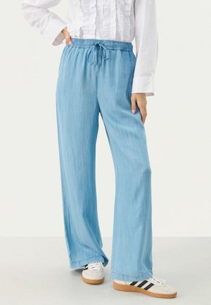 Person trägt hellblaue Hose mit weitem Bein und Kordelzug in der Taille, weiße Rüschenbluse und weiße Sneaker mit schwarzen Streifen.