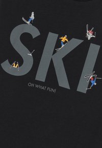 Camiseta negra con la palabra "SKI" en letras negritas grises y ilustraciones estilizadas de esquiadores con trajes coloridos, junto con el texto "¡QUÉ DIVERTIDO!" debajo.