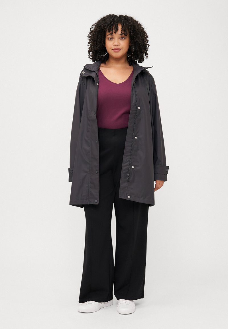 Een vrouw met krullend haar staat zelfverzekerd, gekleed in een donkere regenjas over een bordeaux rood top en zwarte broek, gecombineerd met witte sneakers.