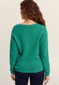 Groene, oversized trui gemaakt van een zachte, gestructureerde stof. Heeft een brede halslijn en geribbelde manchetten, gecombineerd met donkere jeans.